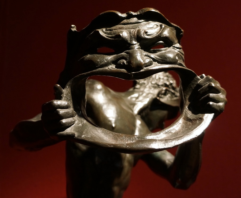 Escultura en bronce de un actor sujetando una máscara con una boca muy grande.