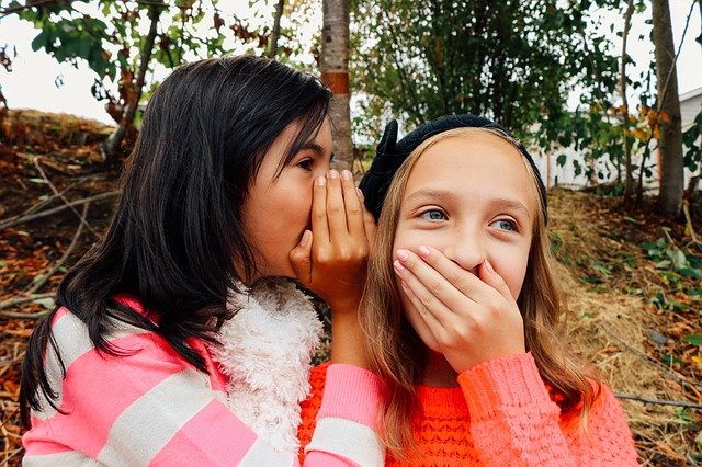 Fotografía de dos niñas en las que una de ellas habla al oído de la otra en acción de contar algo.