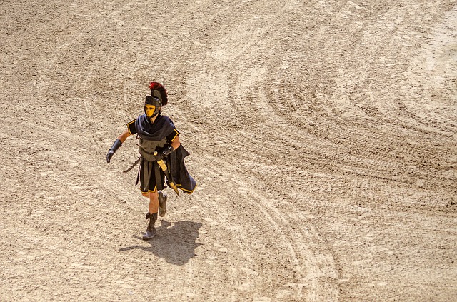 Fotografía de un actor caracterizado como un romano de la época en un lugar con arena representando un duelo de gladiadares.