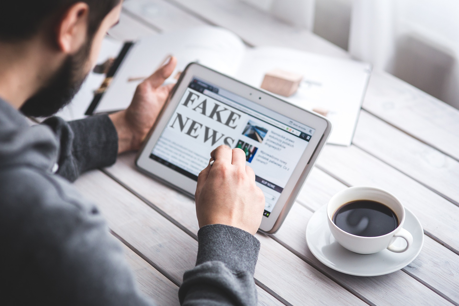 Una fotografía en la que hay un chico leyendo el periódico en una tablet mientras toma un café. En la cabecera de la noticia se puede leer "fake news"