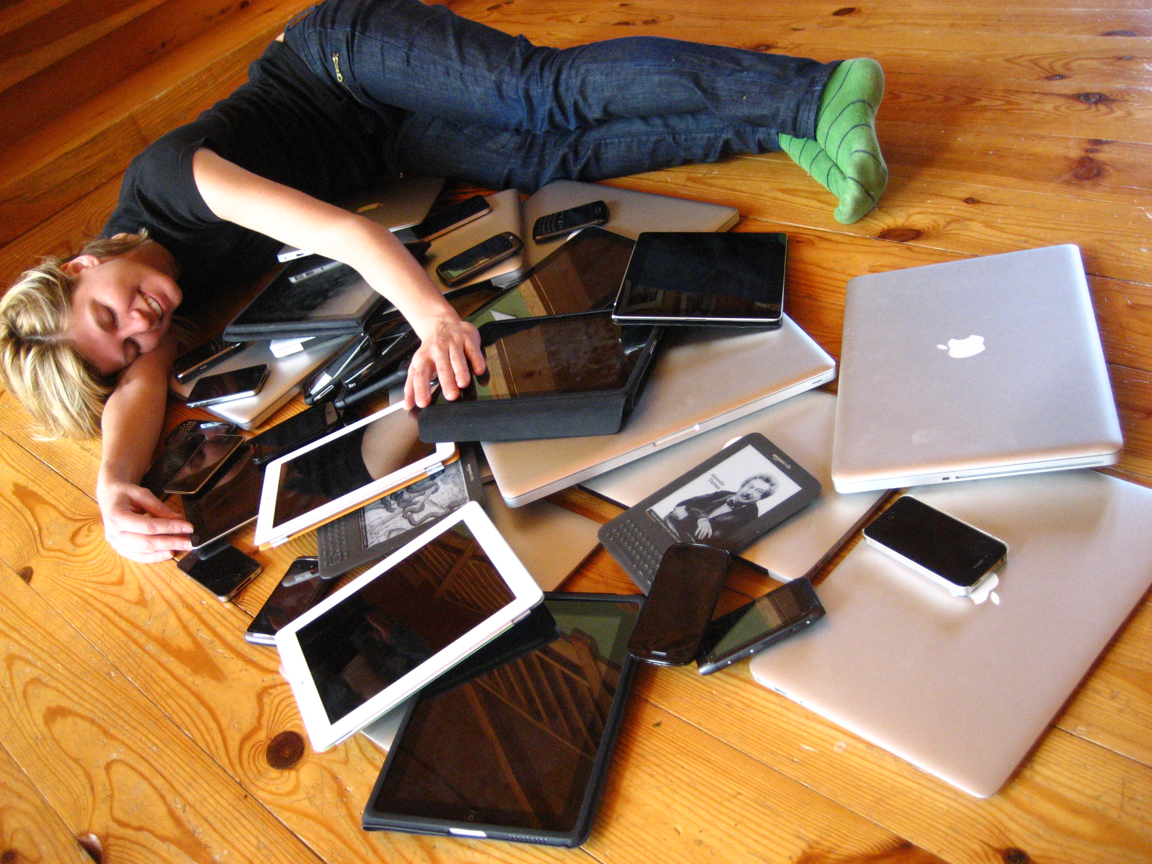 Una persona tumbada en el suelo y rodeada de aparatos electrónicos (teléfonos, ordenadores, etc.).